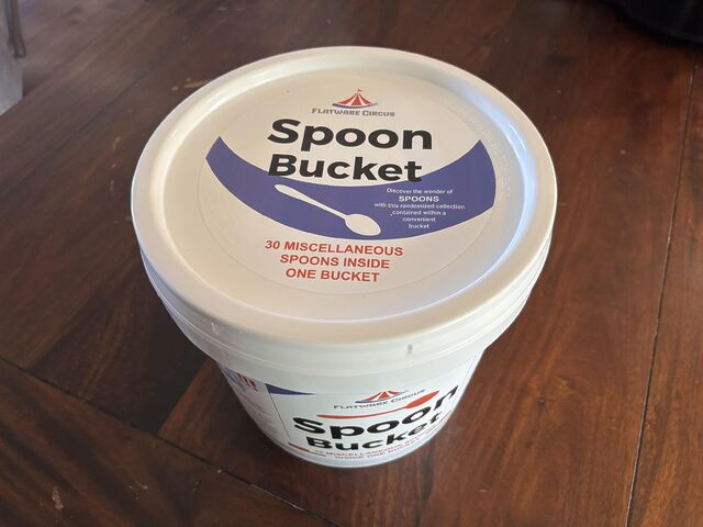 2 gallon white plastic bucket, labeled: - Flatware Circus - Spoon Bucket - Discover the wonder of spoons with this randomized collection contained within a convenient bucket - 30 miscellaneous spoons inside one bucket