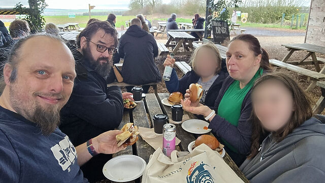 A family of two white men with beards and one white woman sit at a picnic bench in a barn, alongside their two (blurred) children. They're holding baps with sausages, bacon, or eggs in, and surrounded by canned drinks and takeaway coffee cups. A tote bag nearby gives away that they're at the cafe of Diddly Squat Farm.
