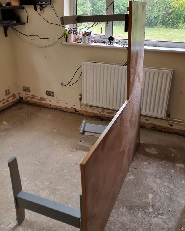 A large, L-shaped office desk with thick motorised metal legs lies on its edge, upright, in a bare concrete-floored home office.