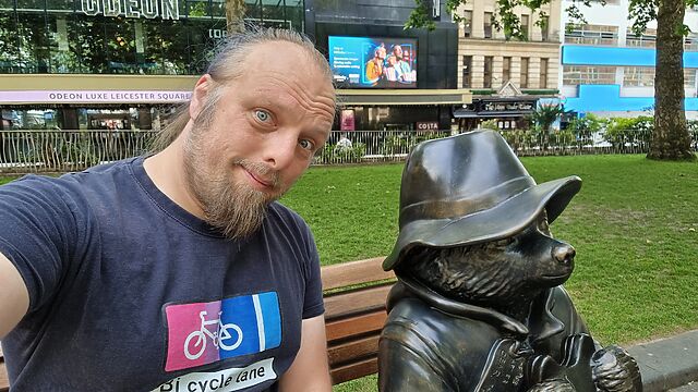 Dan sits on a park bench in Leicester Square, alongside a bronze sculpture of Paddington Bear.