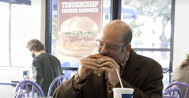 David Cross, as Arrested Development's Tobias Fünke, bites into a burger in a Burger King restaurant, with a Chicken Tendercrisp advertisement prominently displayed in the background.