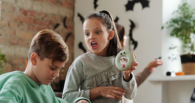 A child with a ring doughnut decorated to look like a monster imitates the face of that monster, towards another child who is pointedly not paying attention.