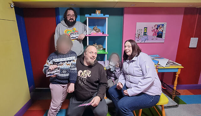 The same adults and children pose in a colourful escape room, with padlocked boxes and banks of light switches visible amongst cat toys.