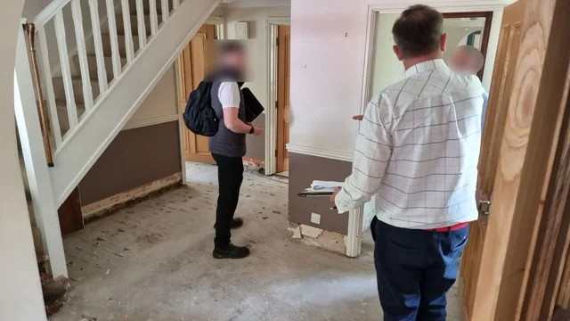 Several men stand on the bare concrete floor of a residential hallway with no furniture or skirting boards.