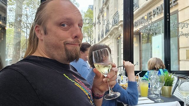 Dan, a white man with a ponytail and a goatee, site in a Parisian cafe sipping a glass of white wine.