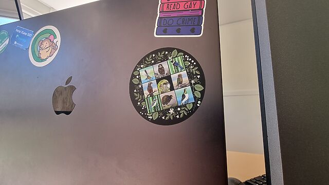 Circular sticker featuring nine New Zealand birds, stuck to a laptop under a sticker that says 'read gay; do crime'.