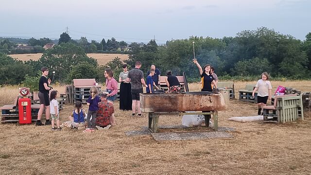 Adults and children mill around a firepit, some of them holding marshmallows on skewers.