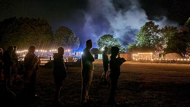 A group of adults and older children stand out in a field at night, while in the distance thick smoke is illuminated by flashing blue lights.