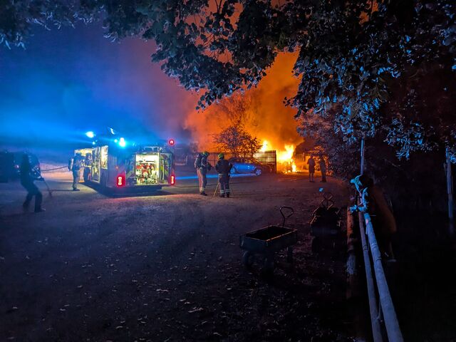 Firefighters pull hoses from a fire engine towards the inferno of a flaming cabin.
