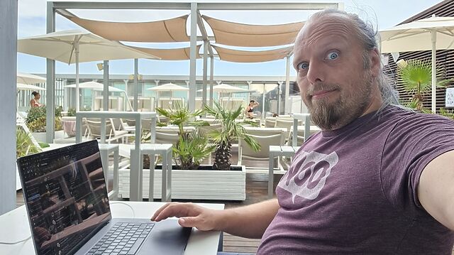 Dan, a white man with a goatee beard and a blue ponytail, sits at a poolside table in a hotel roof terrace, using his laptop alongside a pile of towels.
