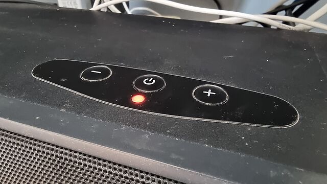 Top surface of a TV soundbar showing plus, minus, and power buttons, and a red LED.