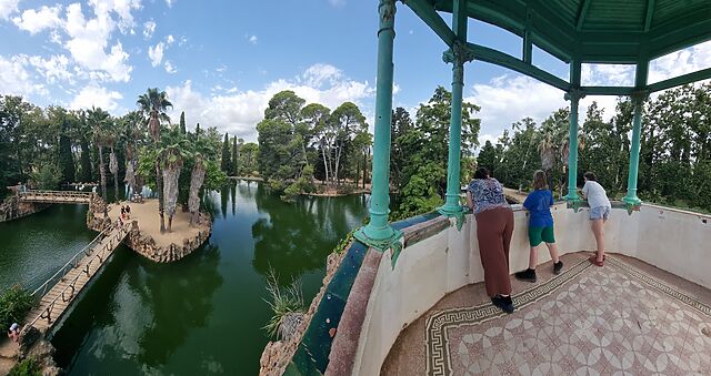Wide/panoramic view from a pagoda-like tower, where a woman and two children lean over a railing to look at a network of bridge-linked islands in a lake below, dotted with palm and other trees.