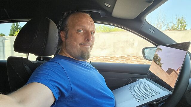 Dan, a white man with a short 'goatee' beard and a blue ponytail, sits in the drivers' seat of a left-hand drive car with a laptop perched between his belly and the steering wheel, giving a sarcastic grin to the camera.