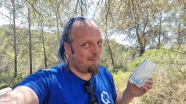 Dan, sweating, holds a medium-sized tupperware tub in a hilly forest.