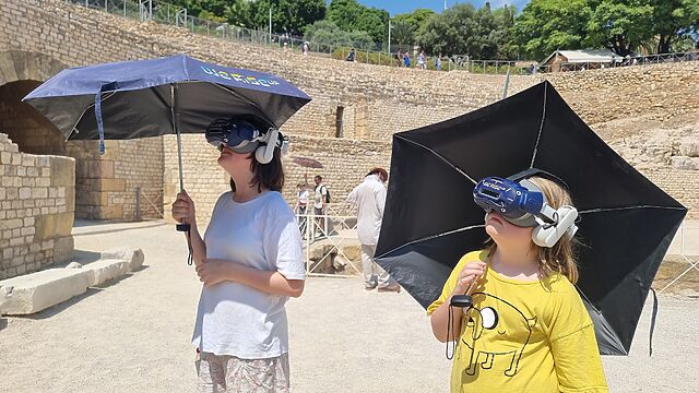 Two children wearing VR headsets and holding parasols, in the ruins of a restored Roman amphitheatre.