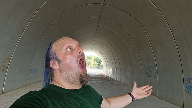 Dan, a white man with a blue ponytail, shouts upwards in a broad concrete tunnel.