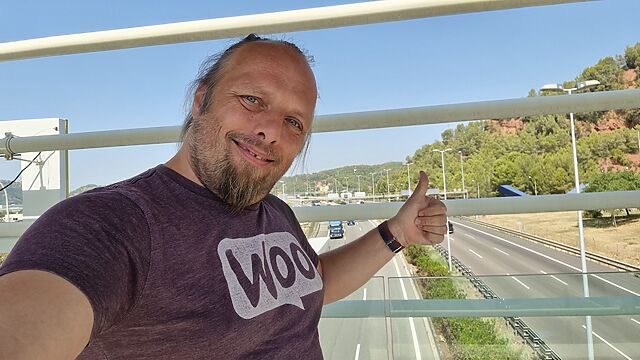 On a semi-closed bridge over a six-lane highway, Dan, wearing a purple 'woo' t-shirt, throws a selfie camera a thumbs-up.