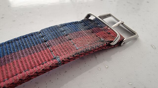 Close up of the buckle end of a worn, grimy watch strap in 'bi pride' colours.