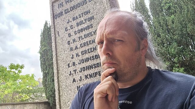 Dan, a white man with a ponytail and goatee beard, sits on a war memorial with a confused expression.