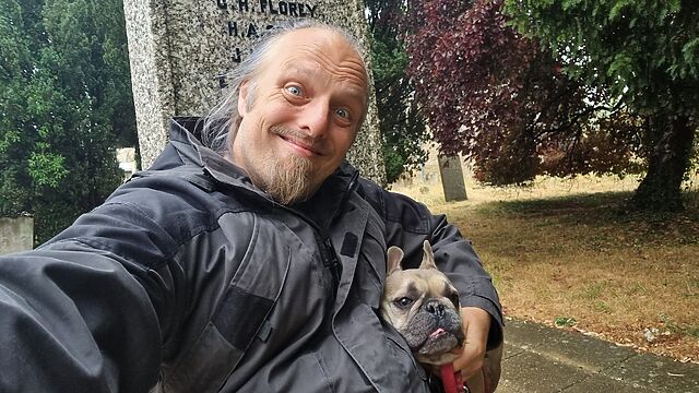 Dan, a white man with a goatee beard, sits on the steps of a rain-wettened village war memorial, hugging a fawn-coloured French Bulldog.