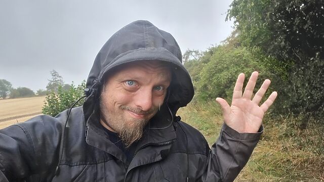 Dan, a white man with a goatee beard, wears a grey hooded jacket on a verdant rural footpath in moderate rain.