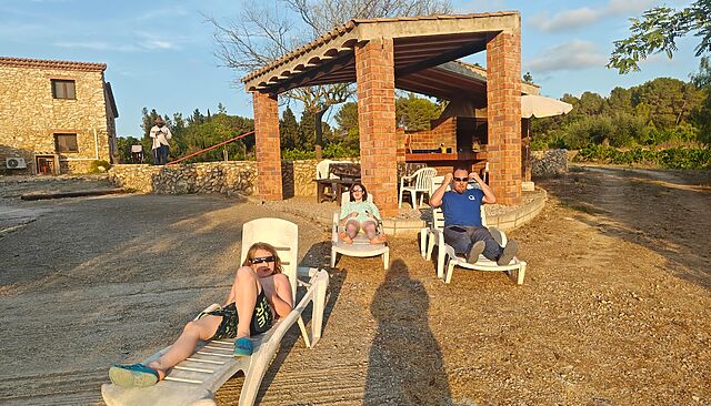 Dan sits on a sun lounger on a driveway, alongside a covered brick barbeque building. He's alongside two children doing the same, and all are wearing disposable eclipse glasses.