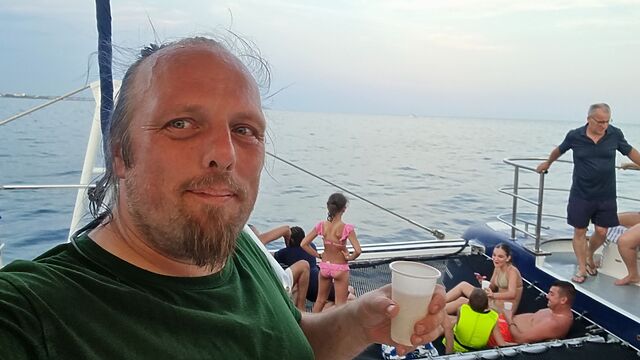 Dan, a white man, drips from a swim in the sea as he stands, holding a plastic cup of white wine, on the deck of a catamaran.