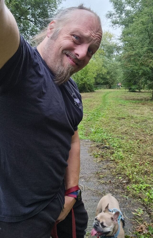 Dan, a white man with a goatee beard, grimaces as he holds his crotch, alongside a French Bulldog on a woodland footpath.