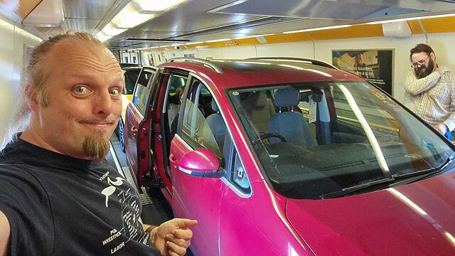 A large red car is parked inside an enclosed car-transporting train. Alongside it Dan, a white man with a goatee beard, takes a selfie.