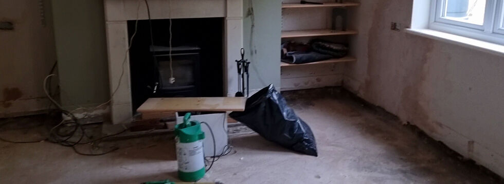 A dilapidated living room with damaged lower walls and missing flooring, filled with tools and bags of rubbish.