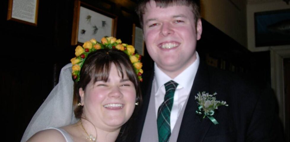 A white couple, bride and groom; she's wearing a white dress and flowers in her hair; he's in a suit with a grey waistcoat and a thistle buttonhole.