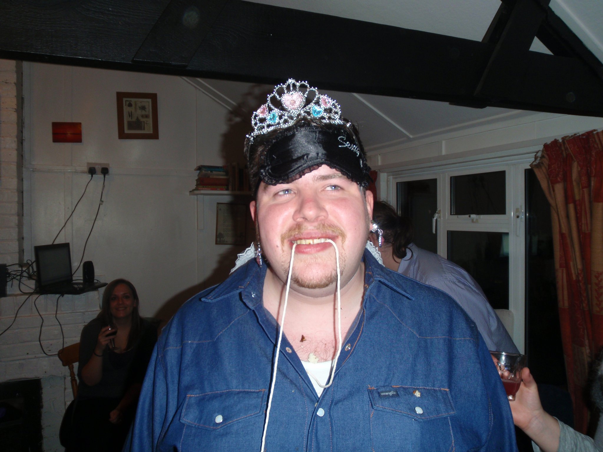 Full image of Alec having lost a doughnut-eating competition; still wearing the blindfold, tiara, and earrings from two previous challenges.