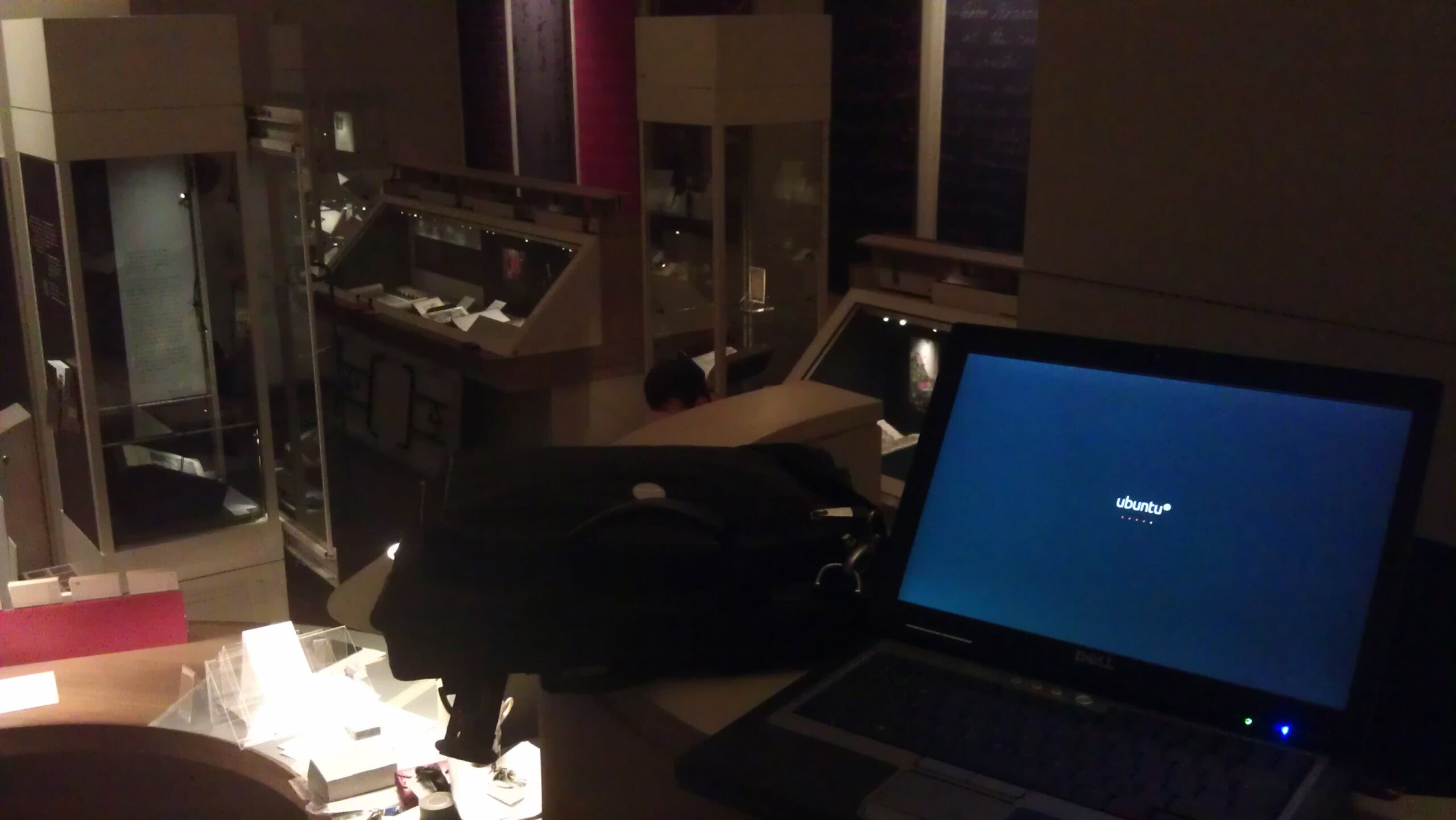 Full image of Photograph showing a laptop running Ubuntu, in front of a partially-constructed exhibition hall in which museum artefacts are being laid-out in glass cases.