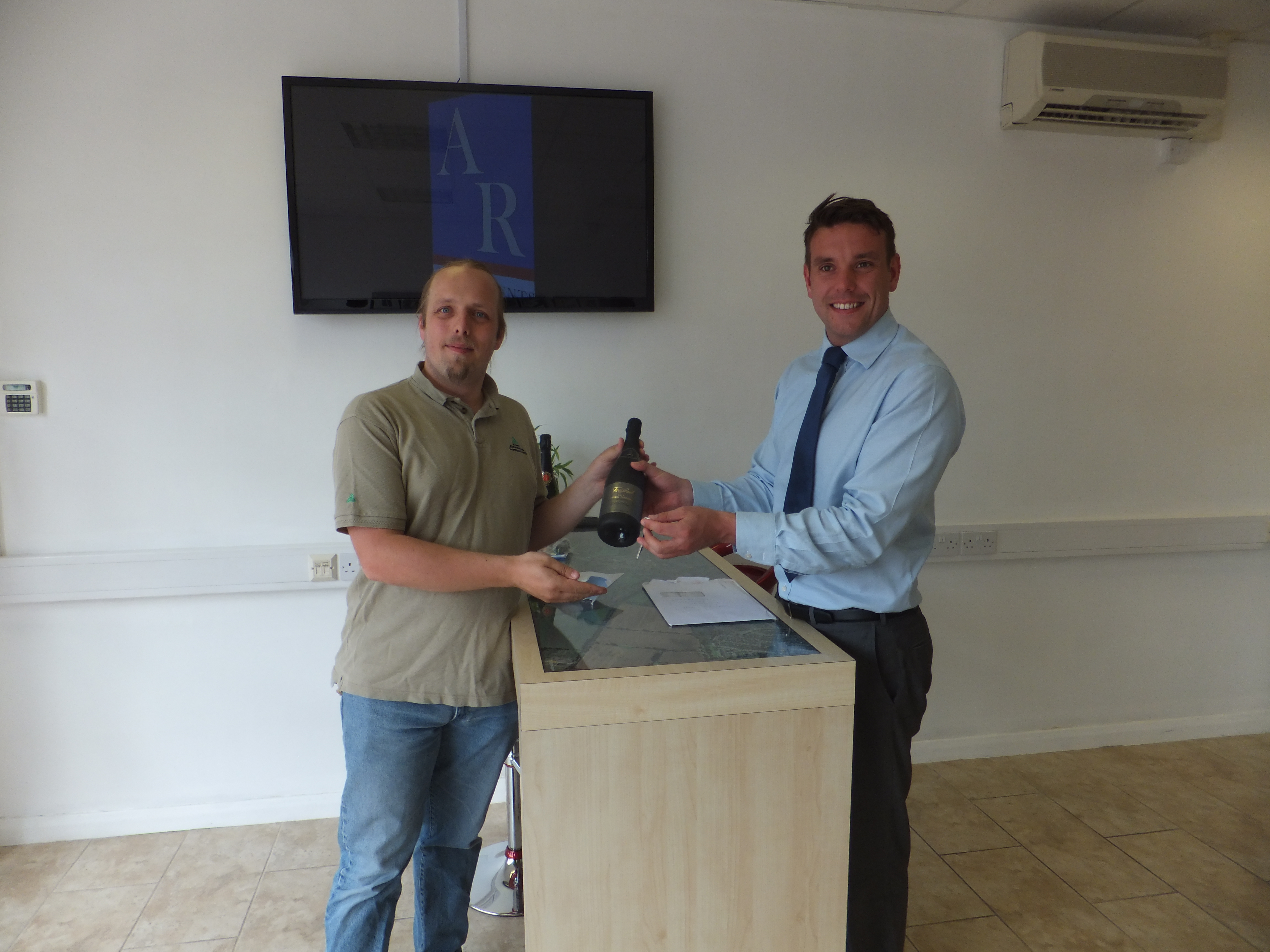 Full image of Dan is handed keys and a bottle of sparkling wine by Mark, the estate agent.