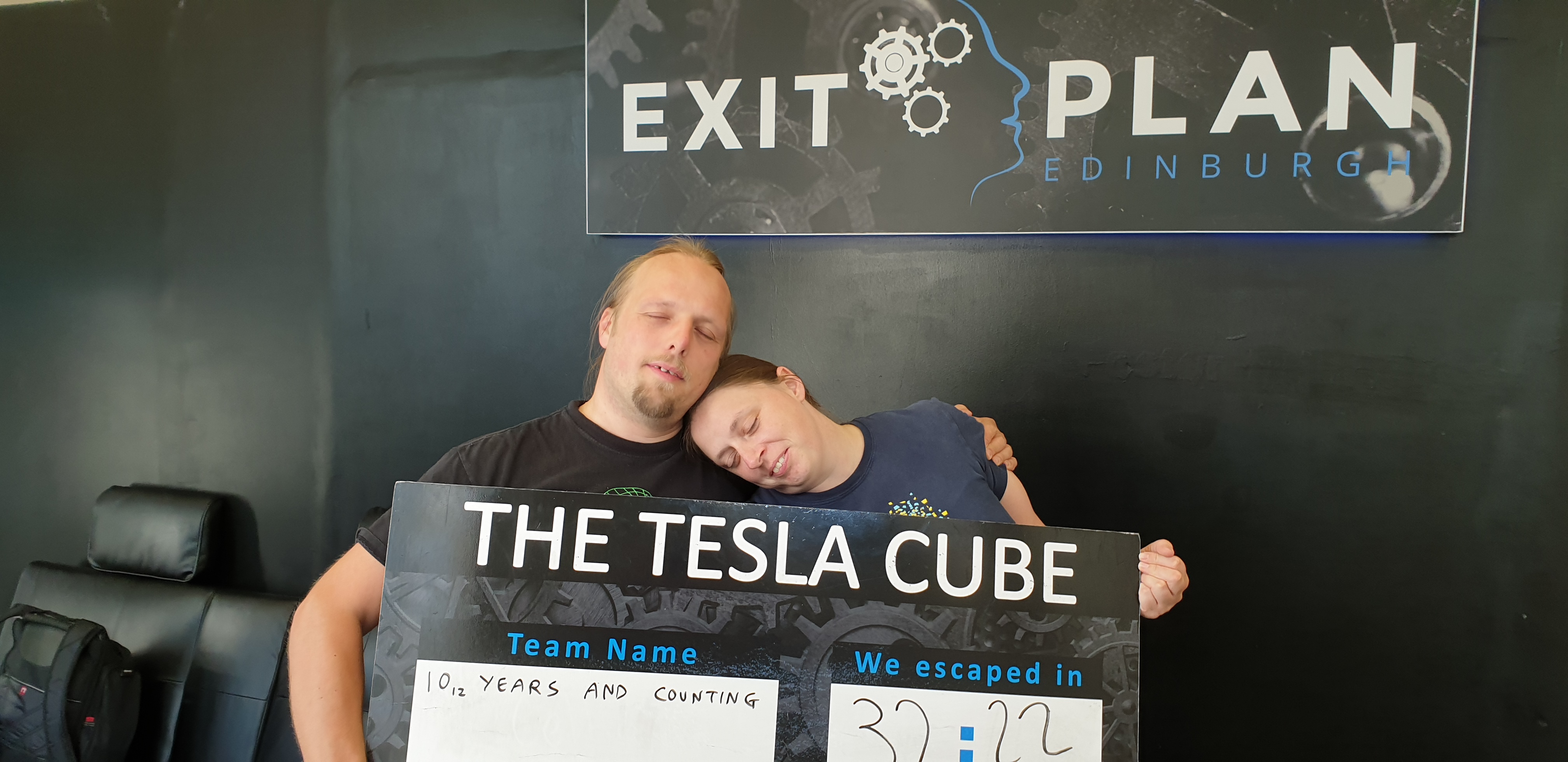 Full image of Dan and Ruth pretend to be asleep while holding a sign that says that they solved The Tesla Cube escape room in 32 mintues and 22 seconds.