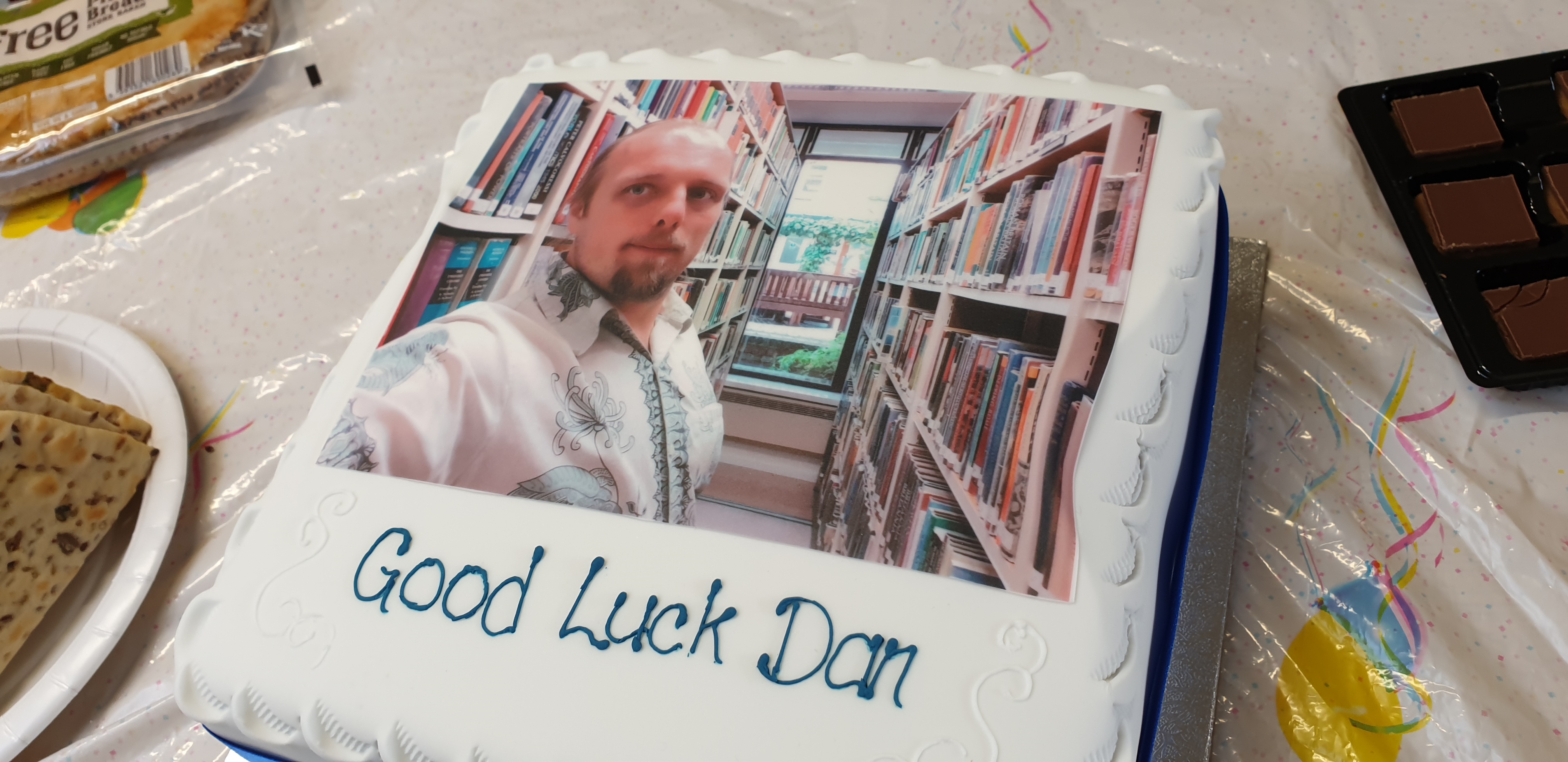 Full image of A cake with icing printed with a picture of Dan in a library. Beneath are iced the words "Good Luck Dan".