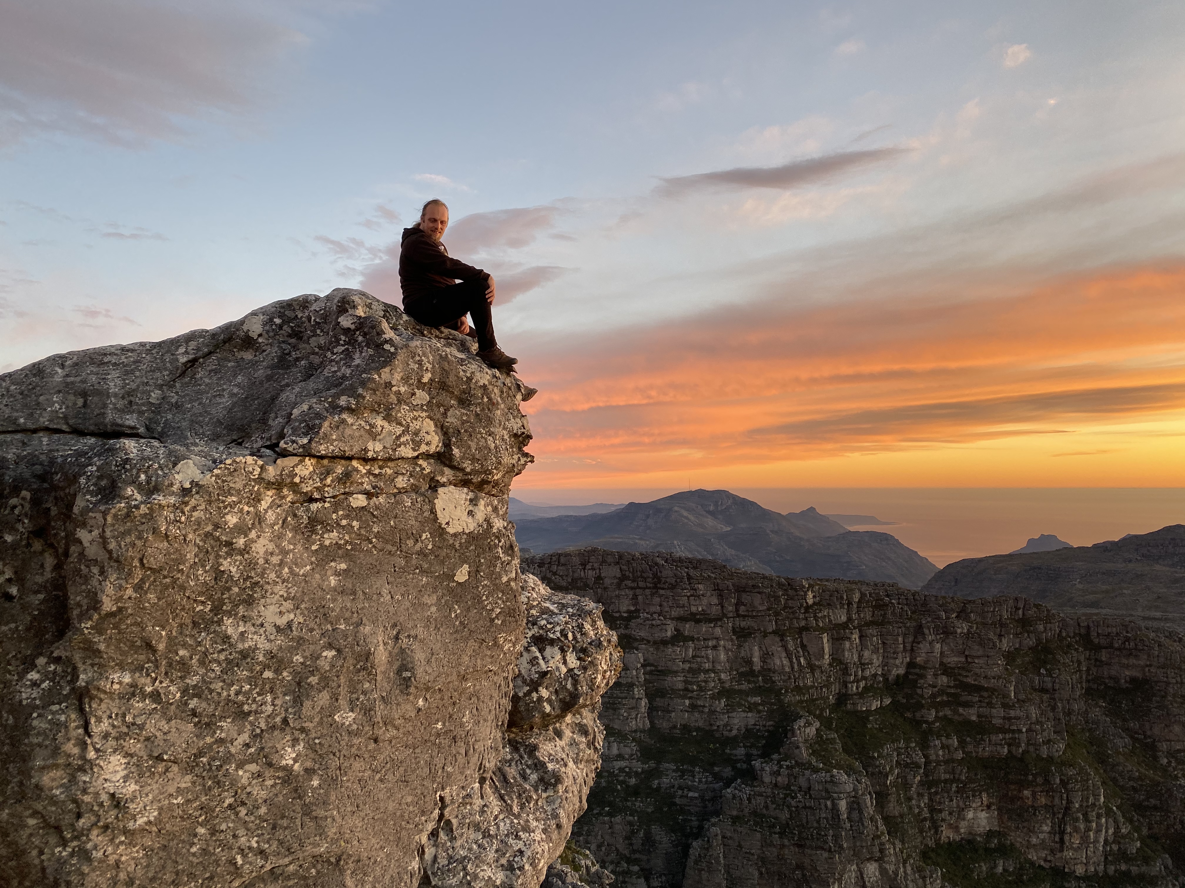 Full image of Dan sitting on a rock on Table Mountain, Cape Town, with a sunset in the background