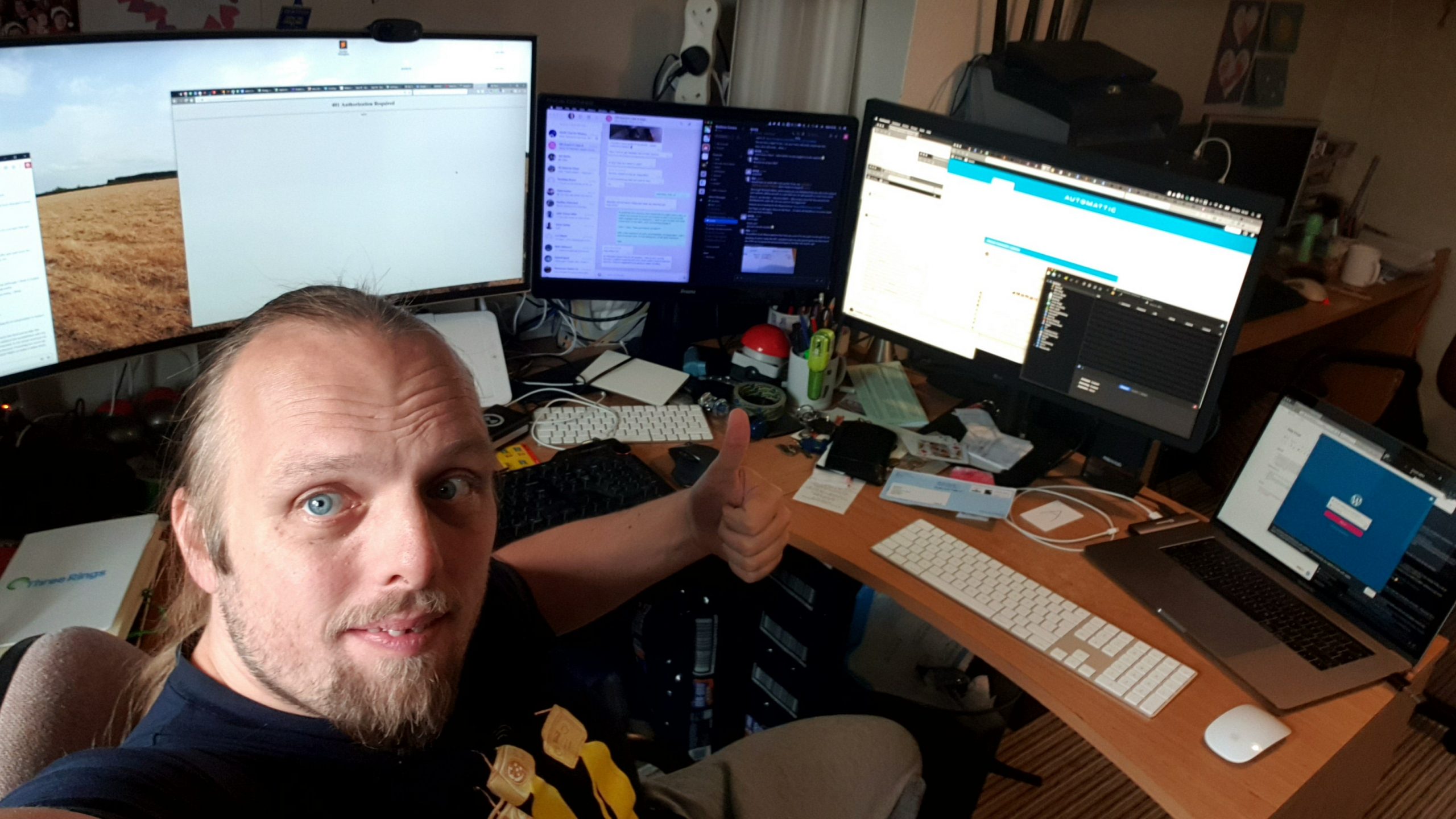 Full image of Dan in front of a crowded desk: two large monitors, a regular-sized monitor, and a laptop are visible, among clutter.