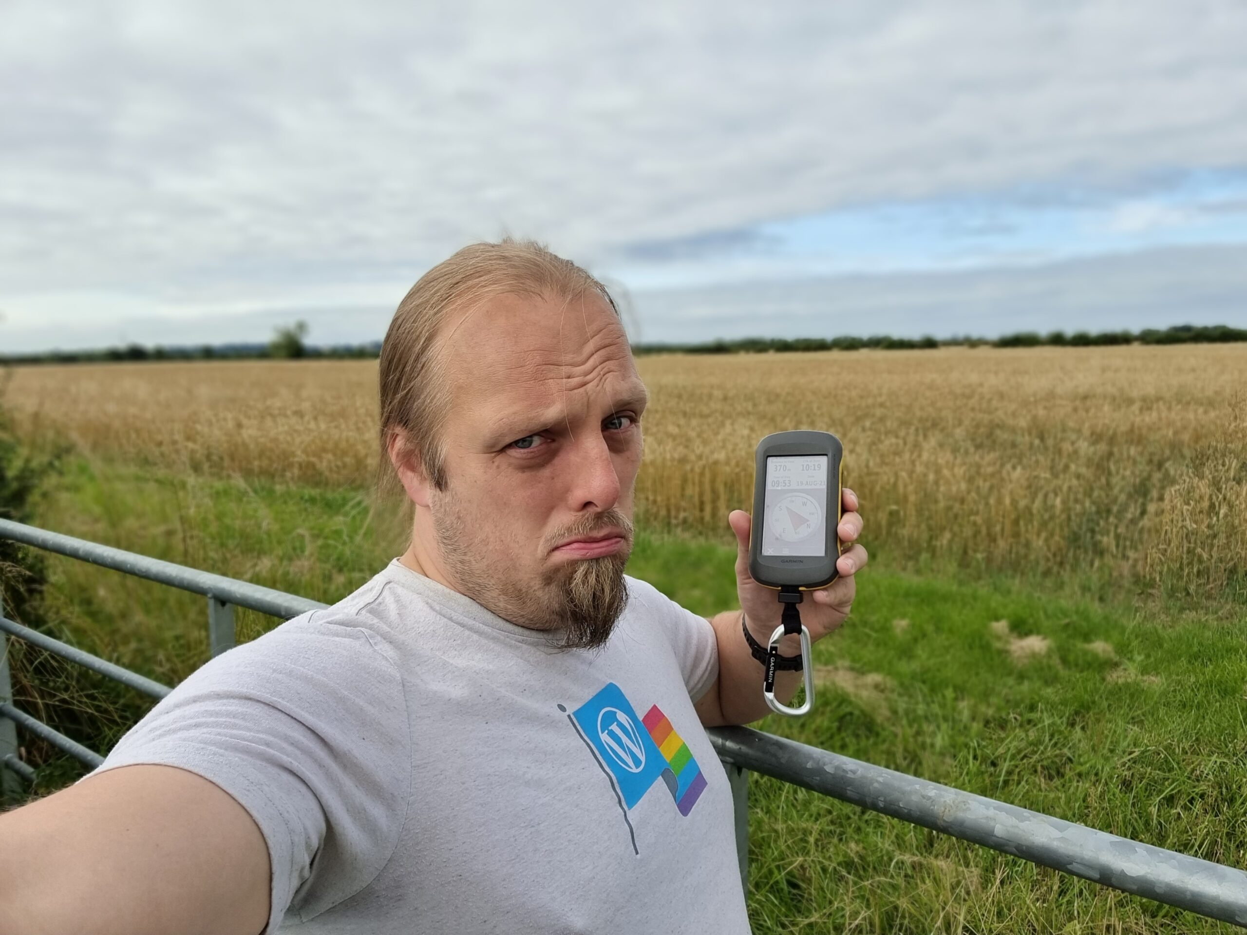 Full image of Dan, looking sad, holding a GPSr at a gate in front of a field of corn.