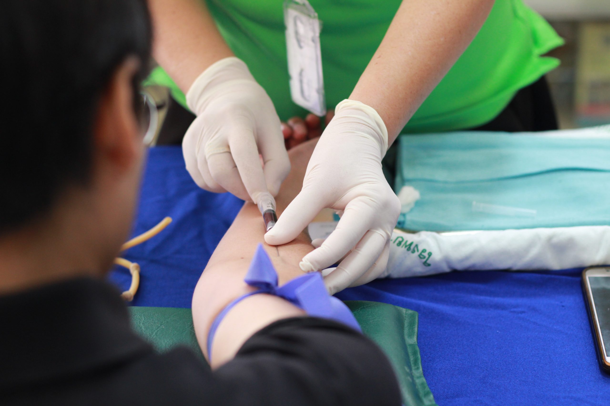 Full image of A nurse wearing gloves uses a hyperdermic needle to take a blood sample from a patients' arm, as seen from over the patient's shoulder.
