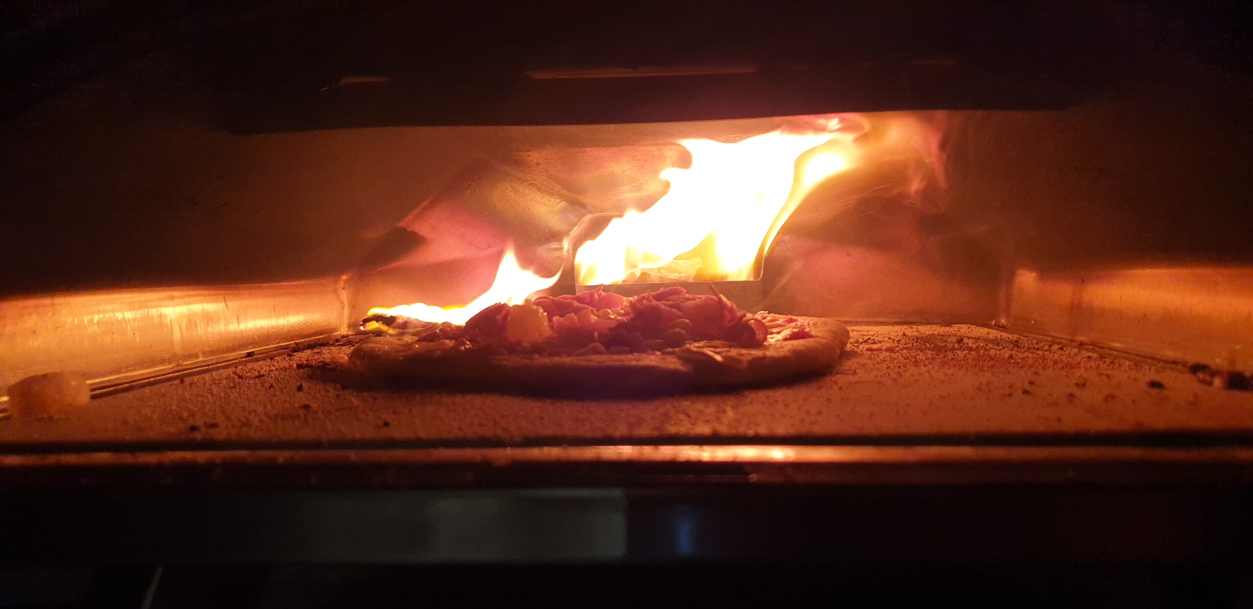 Full image of Pizza in an oven, fire raging behind and jumping onto the floor of the oven.