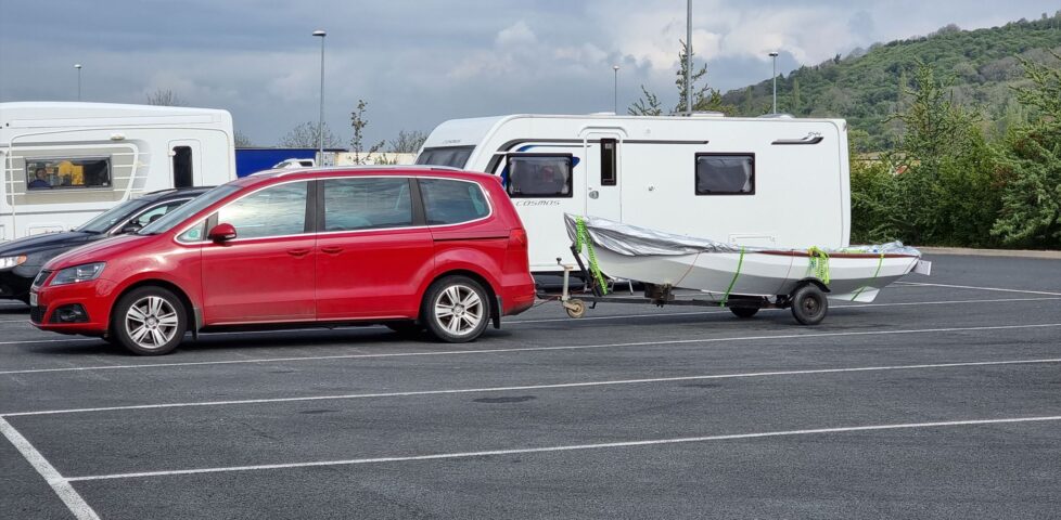 Arthur (red car) and Lucy (rowboat in tow) parked at a service station alongside caravans and HGVs