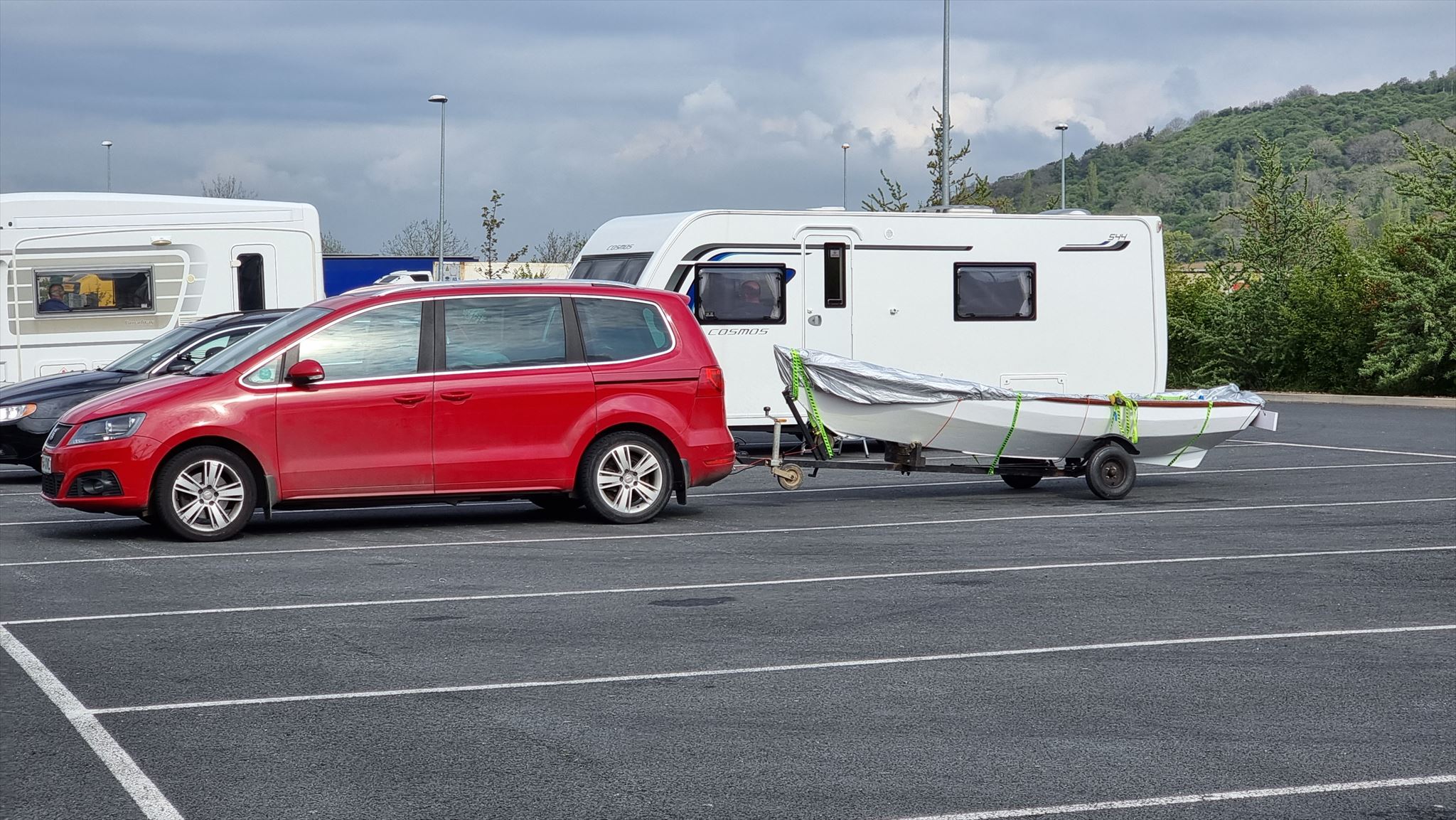Full image of Arthur (red car) and Lucy (rowboat in tow) parked at a service station alongside caravans and HGVs