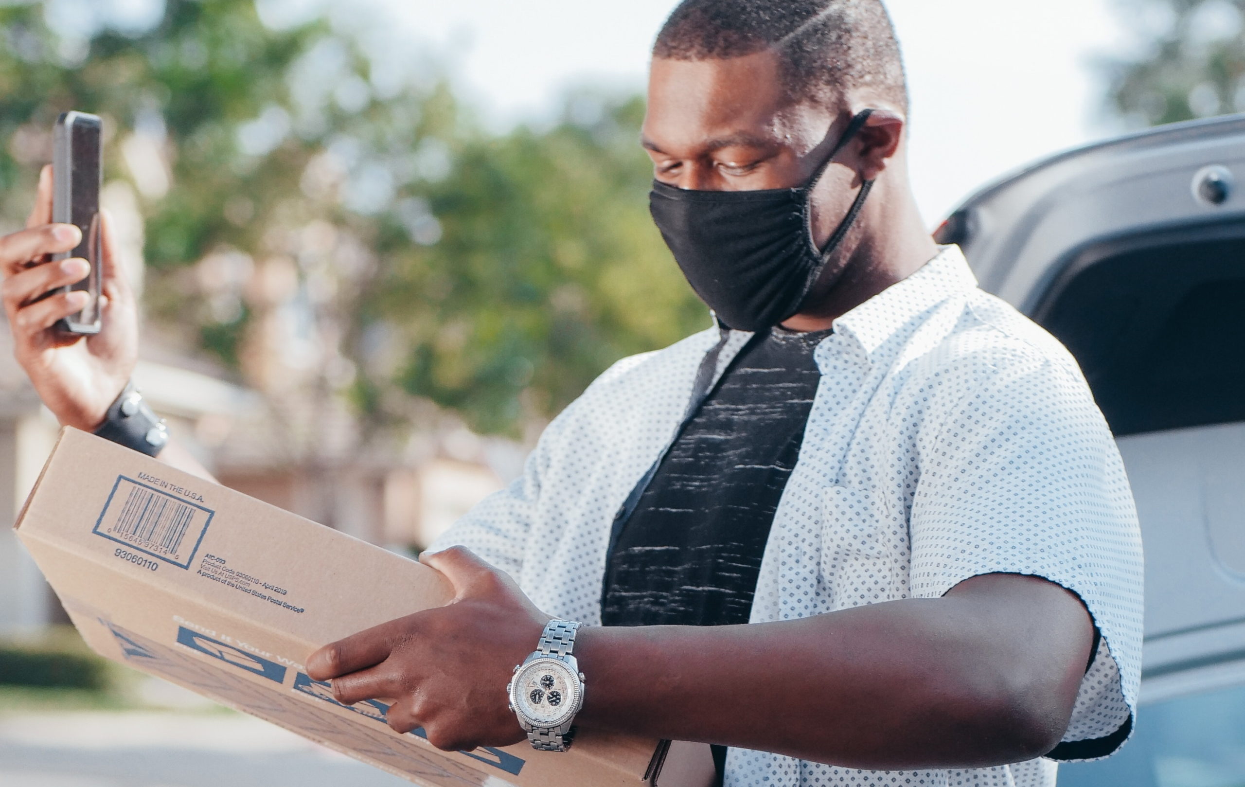 Full image of Delivery man, wearing a face mask, holding a parcel and checking his mobile phone. Photo by Kindel Media from Pexels.