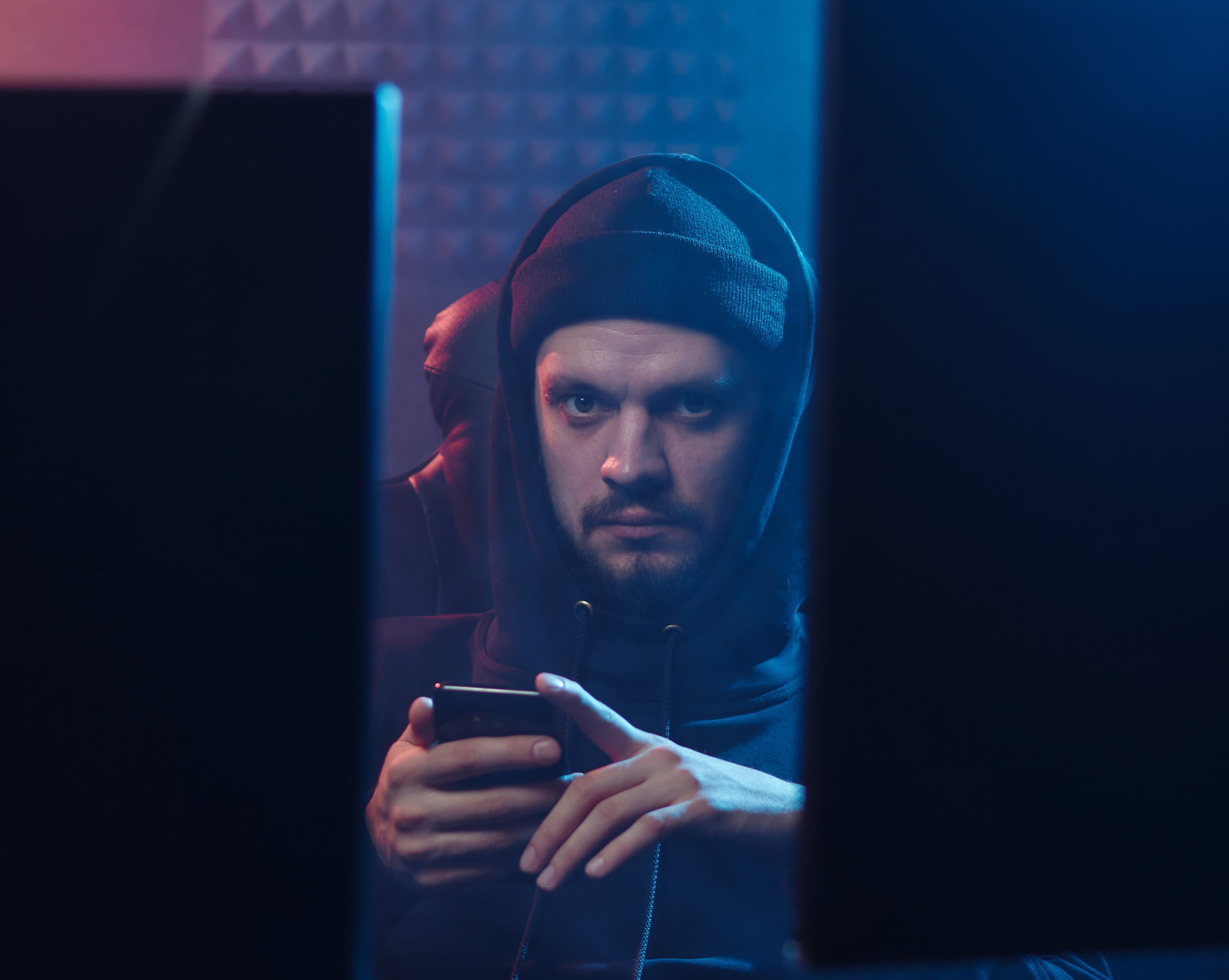 Full image of "Hacker type" man in hoodie between two computer monitors, looking at his phone.