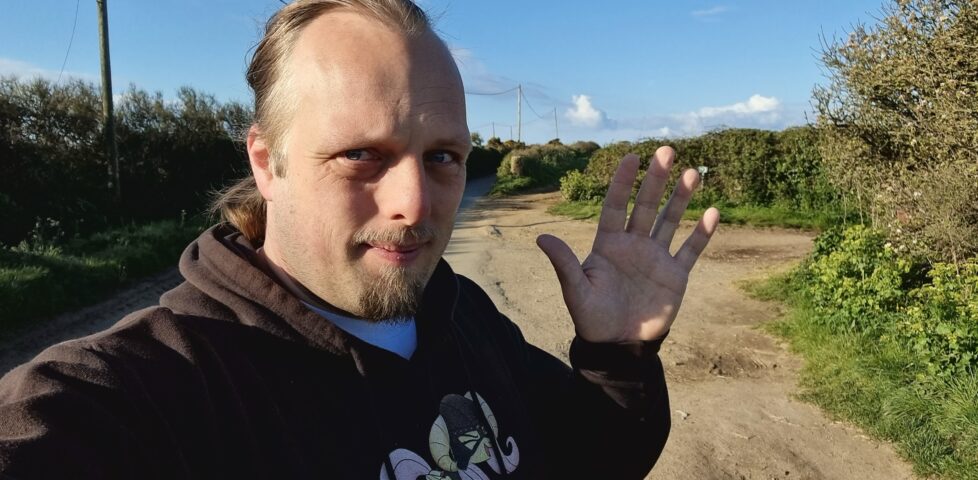Dan, on a country lane in Cornwall, in front of a bright blue sky, waves to the camera.
