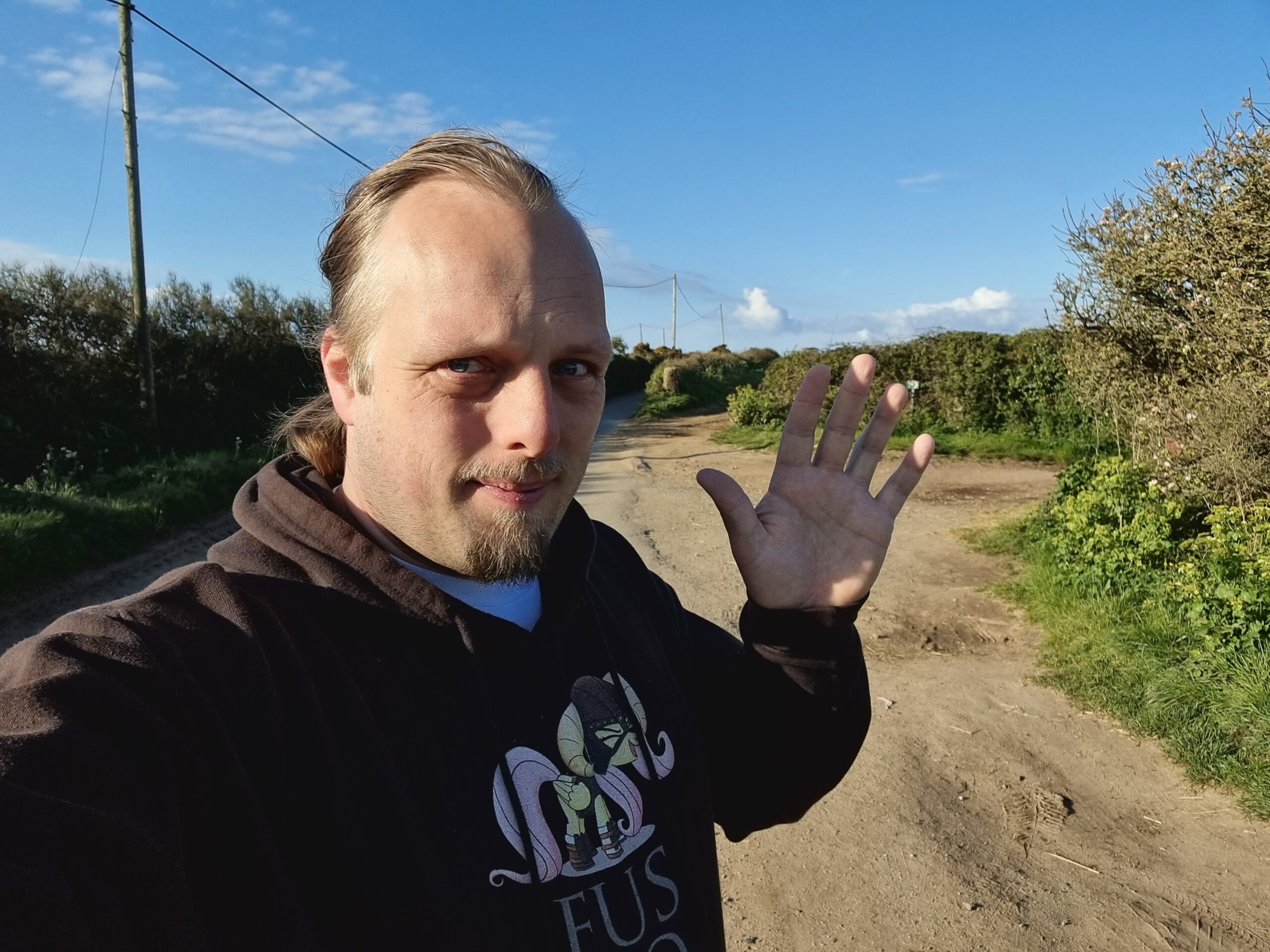Full image of Dan, on a country lane in Cornwall, in front of a bright blue sky, waves to the camera.