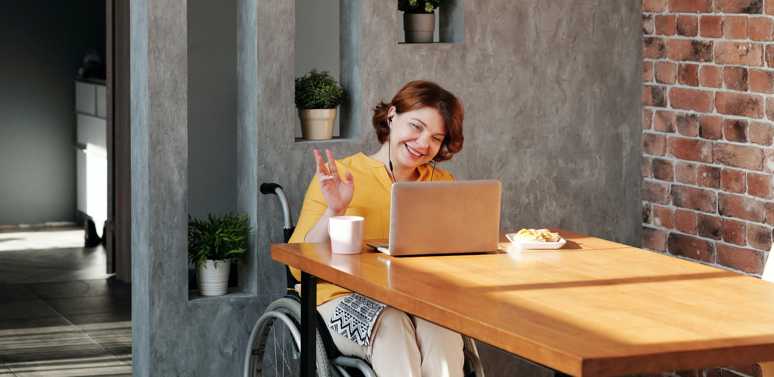 Full image of A woman in a wheelchair waves to a colleague via her laptop screen; she's smiling and has a cup of coffee by her side. Photo by Marcus Aurelius from Pexels.
