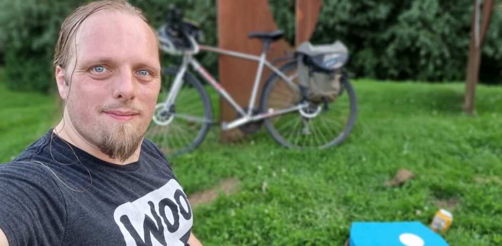 Dan sits on the grass with a Dominos pizza box and a can of beer. His bike is in the background, leaning on a stylistically-corroded piece of metal artwork.