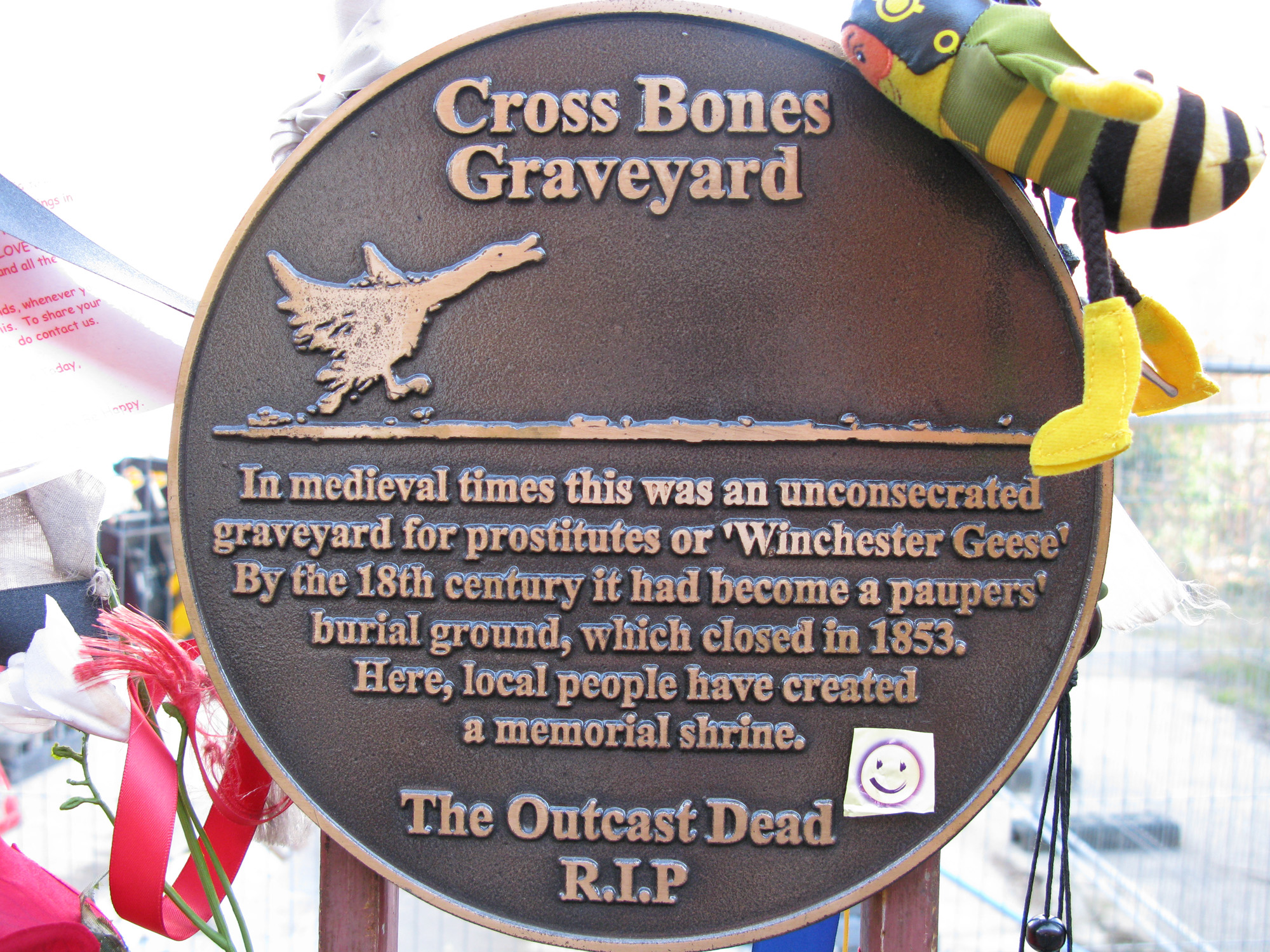 Full image of Plaque with a picture of a goose running and text: "Cross Bones Graveyard. In medieval times this was an unconsecreated graveyard for prostitutes of 'Winchester Geese'. By the 18th century it had become a paupers burial ground, which closed in 1853. Here, local people have created a memorial shrine. The Outcast Dead R.I.P." A smiley face sticker has been attached to the plaque and ribbons and silk flowers are tied nearby.